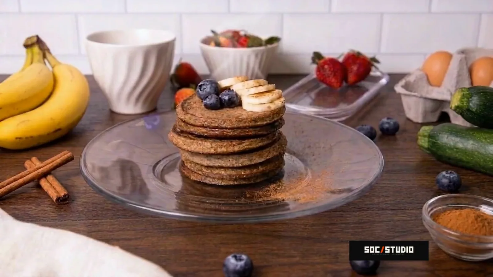 CG food animation still of a pancake stack with banana and blueberries on a glass plate, on a wooden table